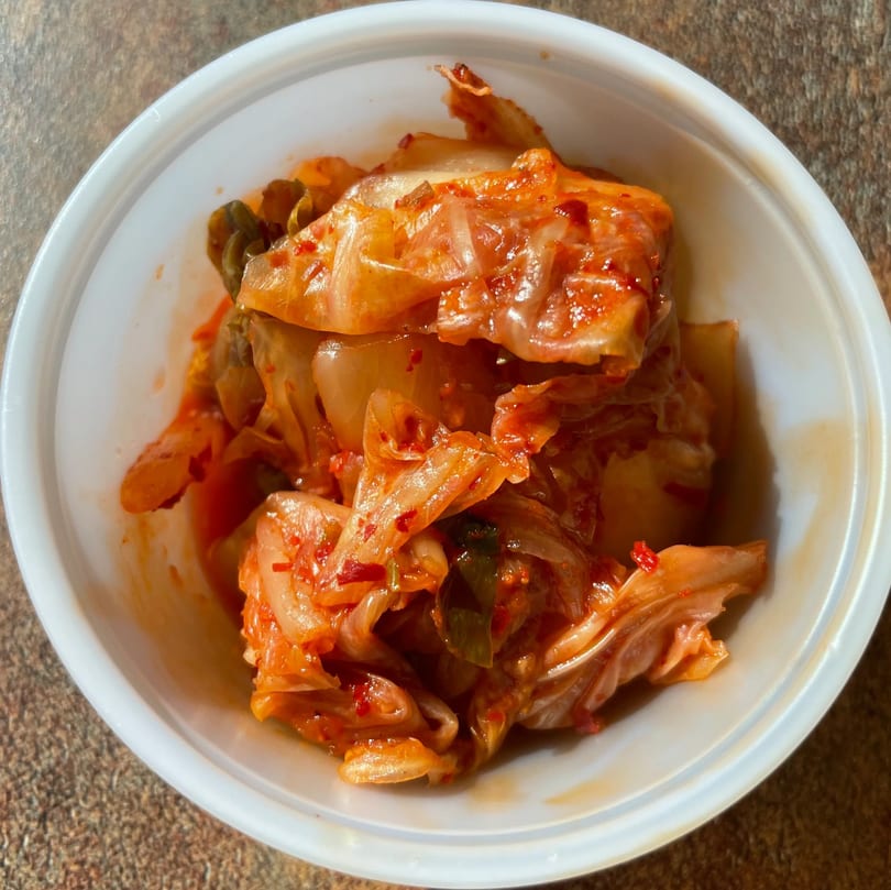 A white bowl filled with vibrant red kimchi, a traditional Korean side dish from Sue's Kimbap House.